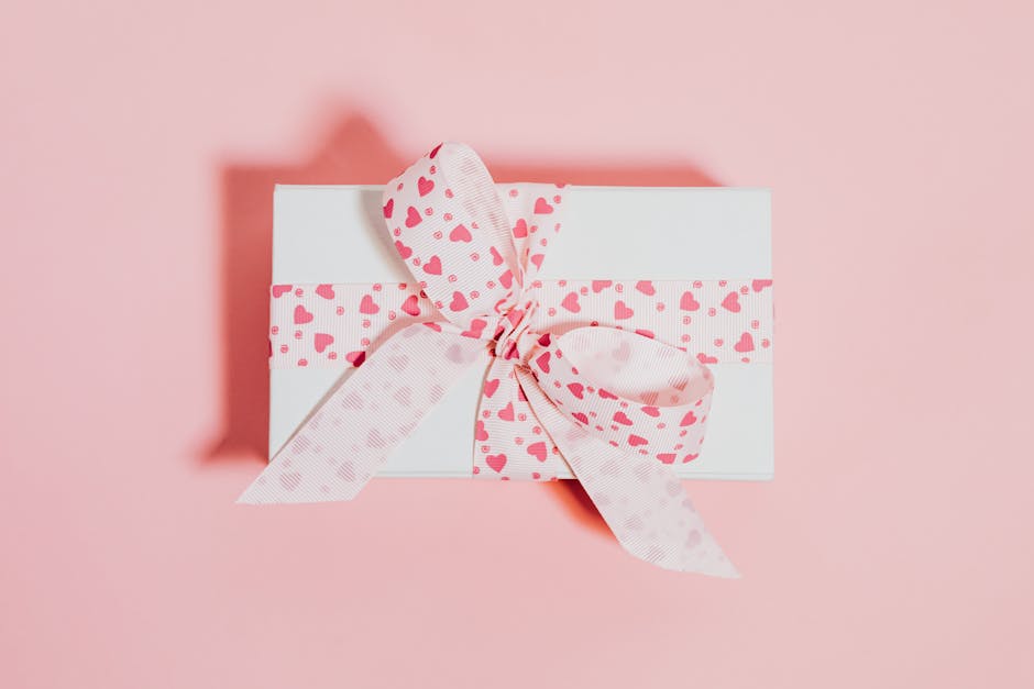 White gift box with pink heart-patterned ribbon on a soft pink background, ideal for celebrations.