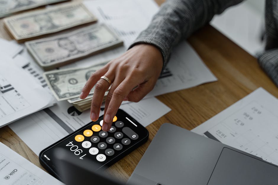 Close-up of a person using a smartphone calculator amid money and financial documents on a wooden table.