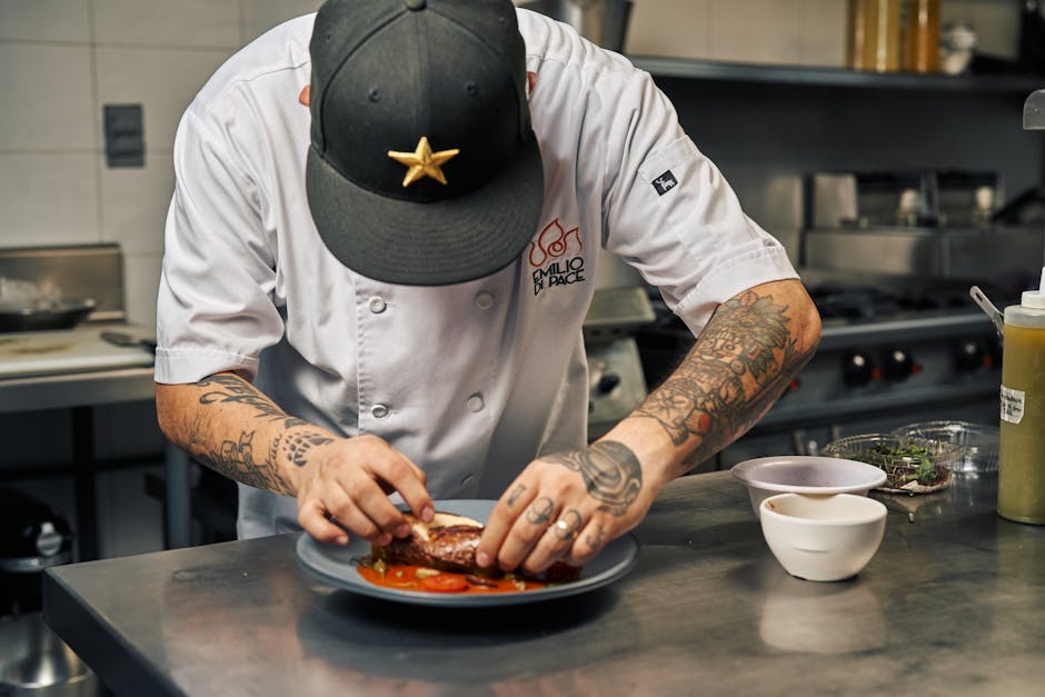 Tattooed chef carefully plates a gourmet dish in professional kitchen setting.