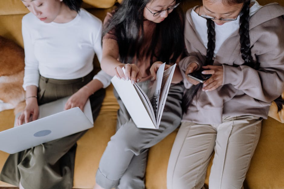 Three women relaxing on a sofa, using gadgets and reading together.