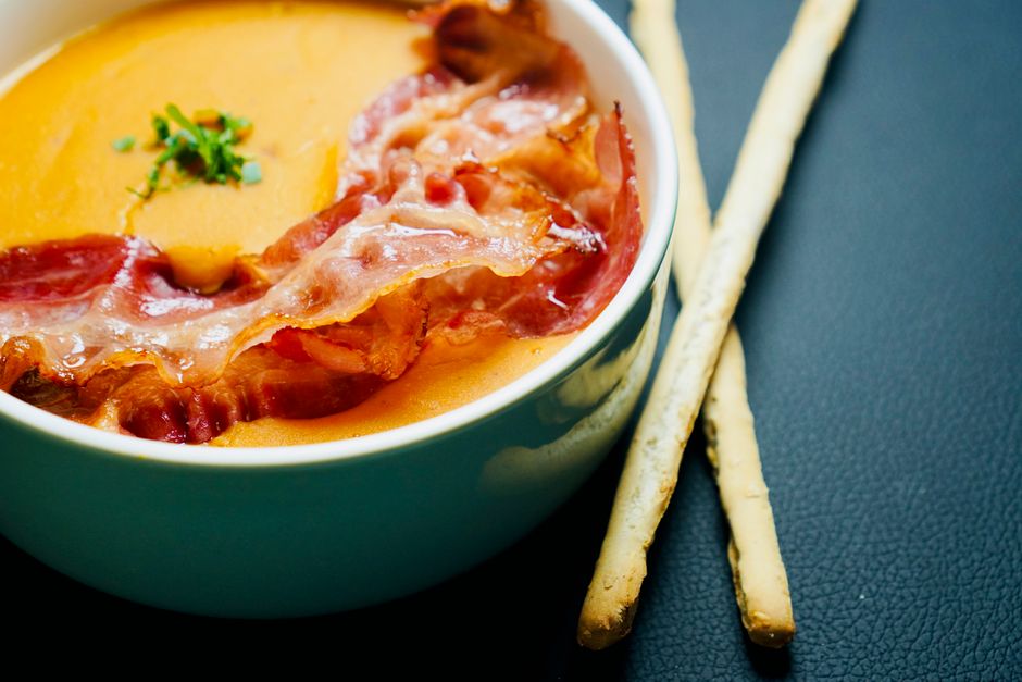 Delicious bowl of creamy soup topped with crispy bacon served with breadsticks on a dark background.