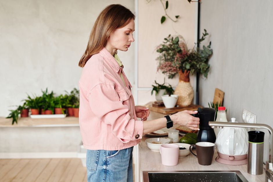 Woman in pink jacket making coffee in a stylish kitchen with plants and decor elements.
