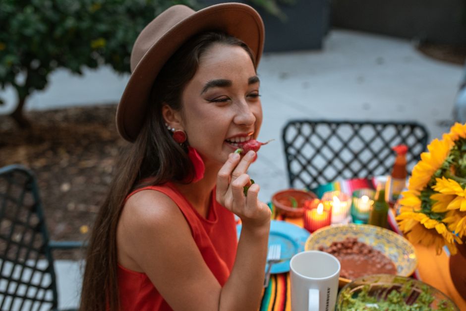 Side view smiling young ethnic female in stylish hat sitting at table during dinner and enjoying ripe radish