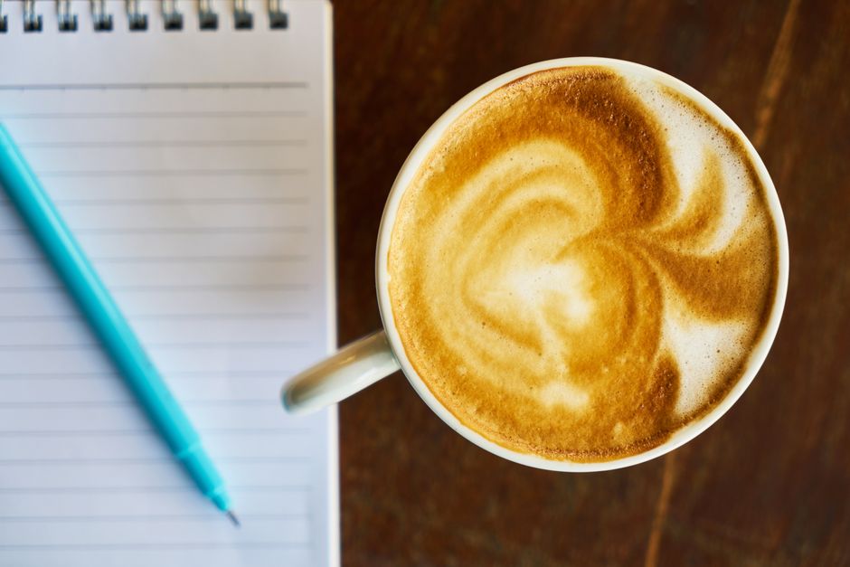 A top view of a frothy latte with artistic foam beside a notepad and pen on a wooden table.