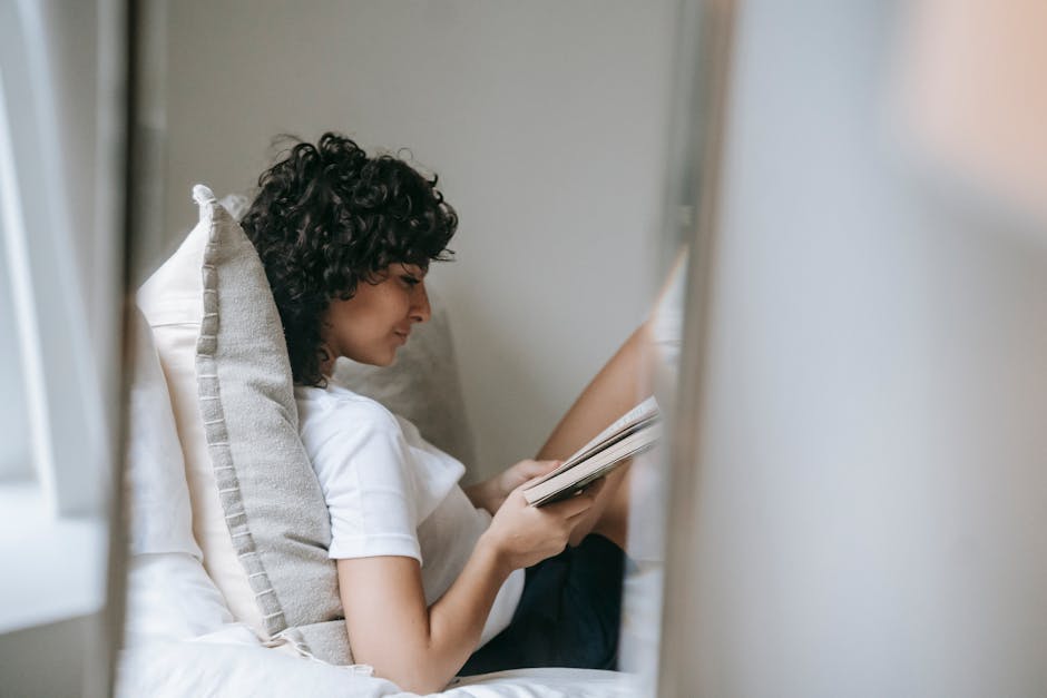 Side view of young curly haired female in sleepwear relaxing on soft bed with pillows while reading paper book