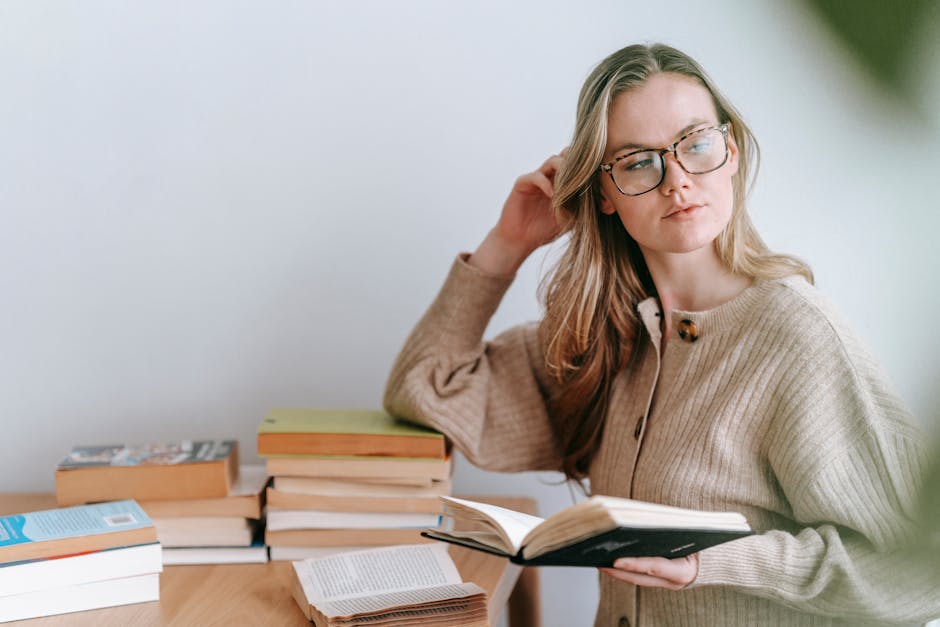 Pensive female student with blond hair in eyeglasses and casual clothes standing with book and looking away against light wall in room