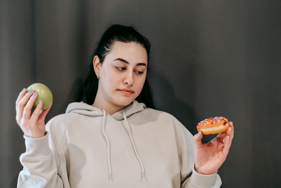 Contemplative plump female in hoodie choosing between ripe green apple and delicious donut in studio